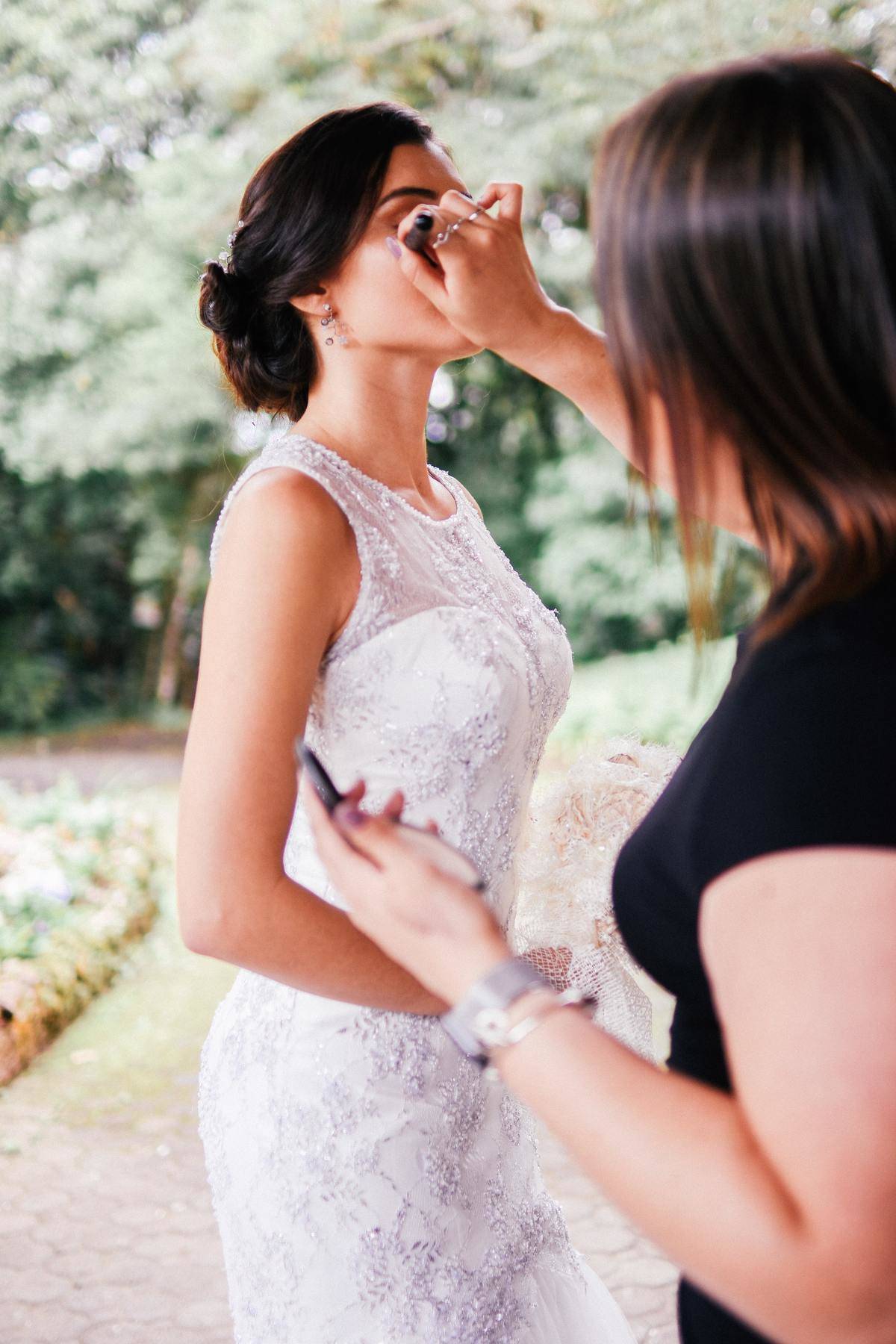 Bride in lace tank top style gown getting makeup touched up by woman with brown hair.
