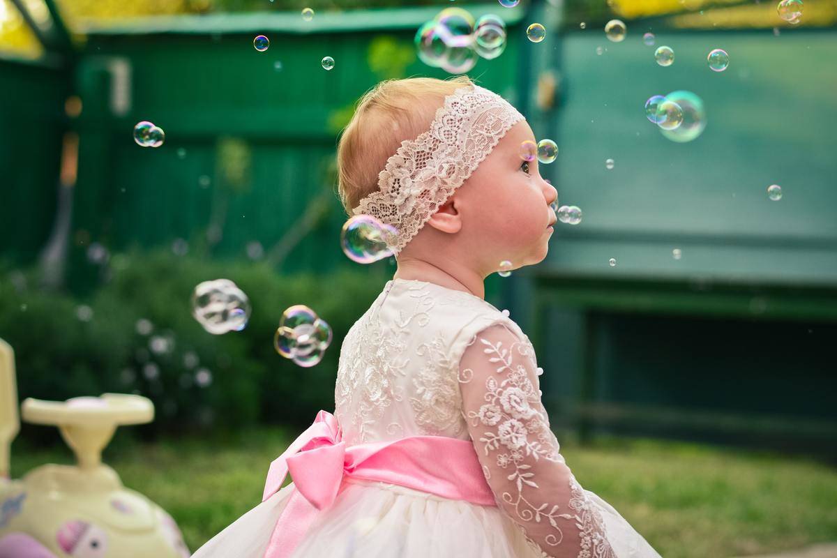 Image of small toddler girl wearing a vanilla-colored lace dress and matching lace headband, a pink sash around middle. Bubbles blowing in air.