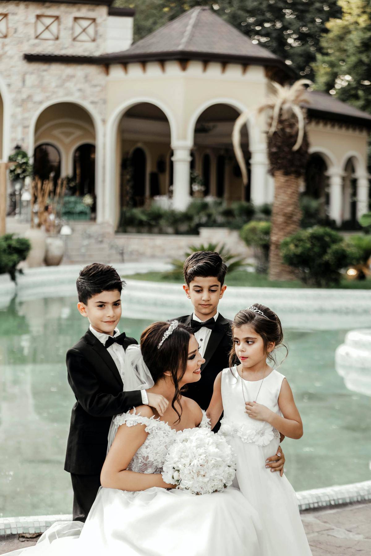 Two young boys in tuxedos stand behind bride who is crouching behind young flower girl. Posing in front of fountain