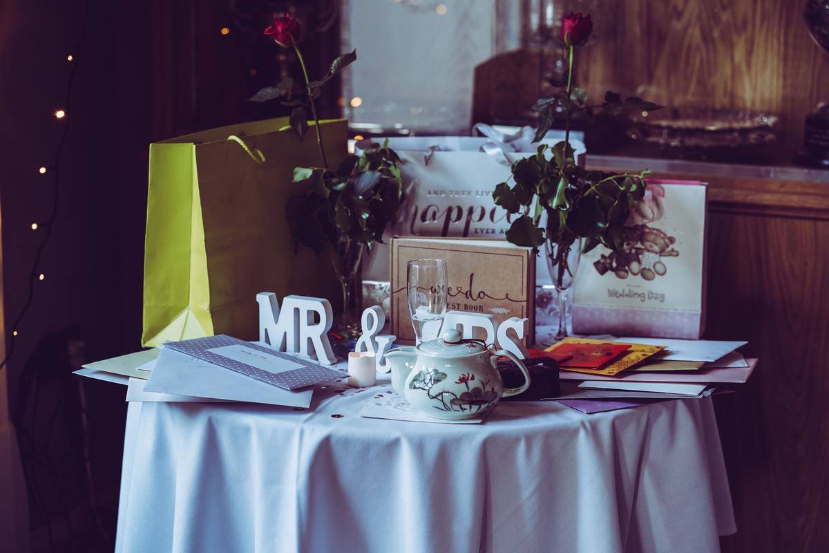 Mr & Mrs. wooden decal signs on a wedding gift table strewn with envelopes. Multiple colorful present bags. White table cloth.