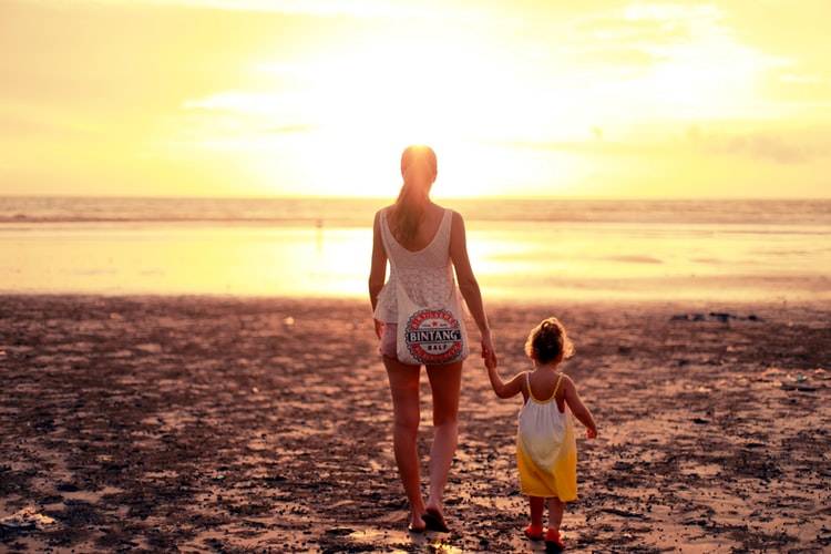 woman holding a younger kid's hand as they walk off into sunset on beach