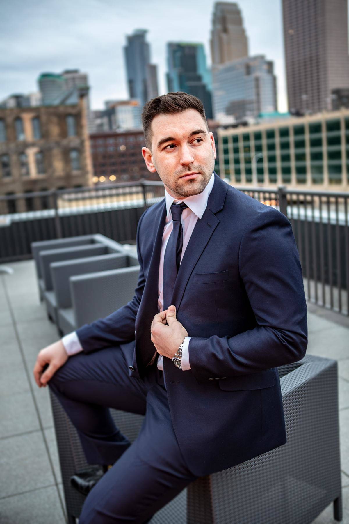 Man in navy blue suit sits on side of chair on rooftop with city skyline behind him