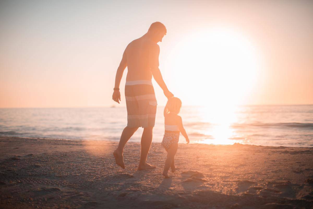 Dad holds small toddler's hand as the sun sets and they walk along the beach