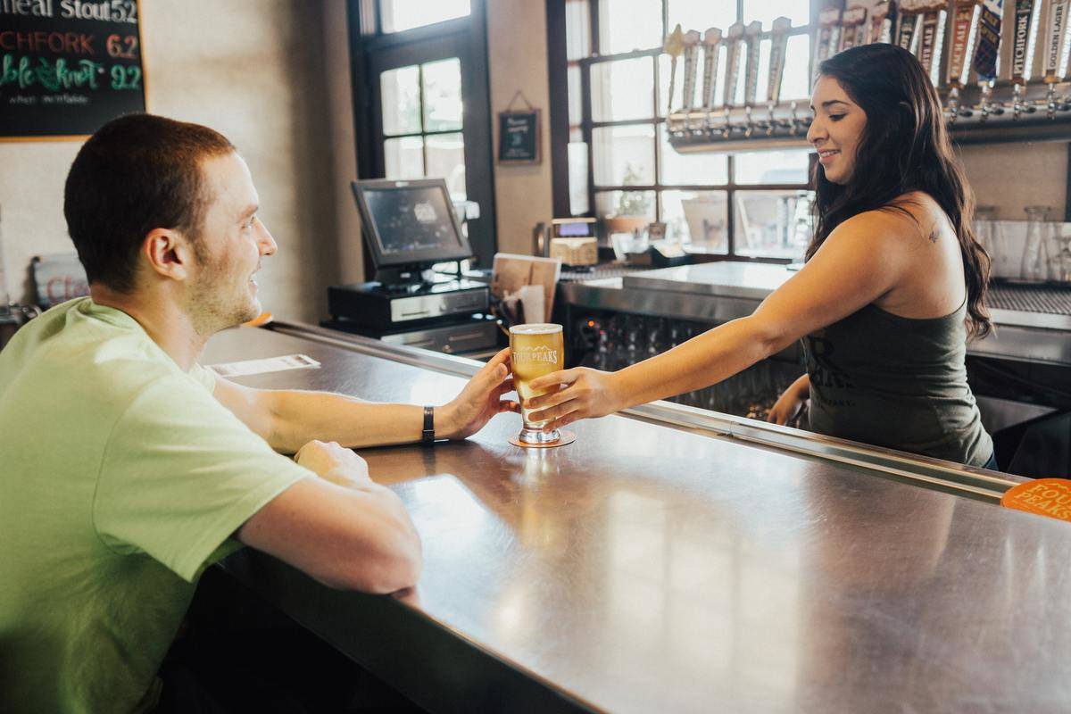 Female bartender passes pint of beer to man sitting across bar, he smiles, she smiles