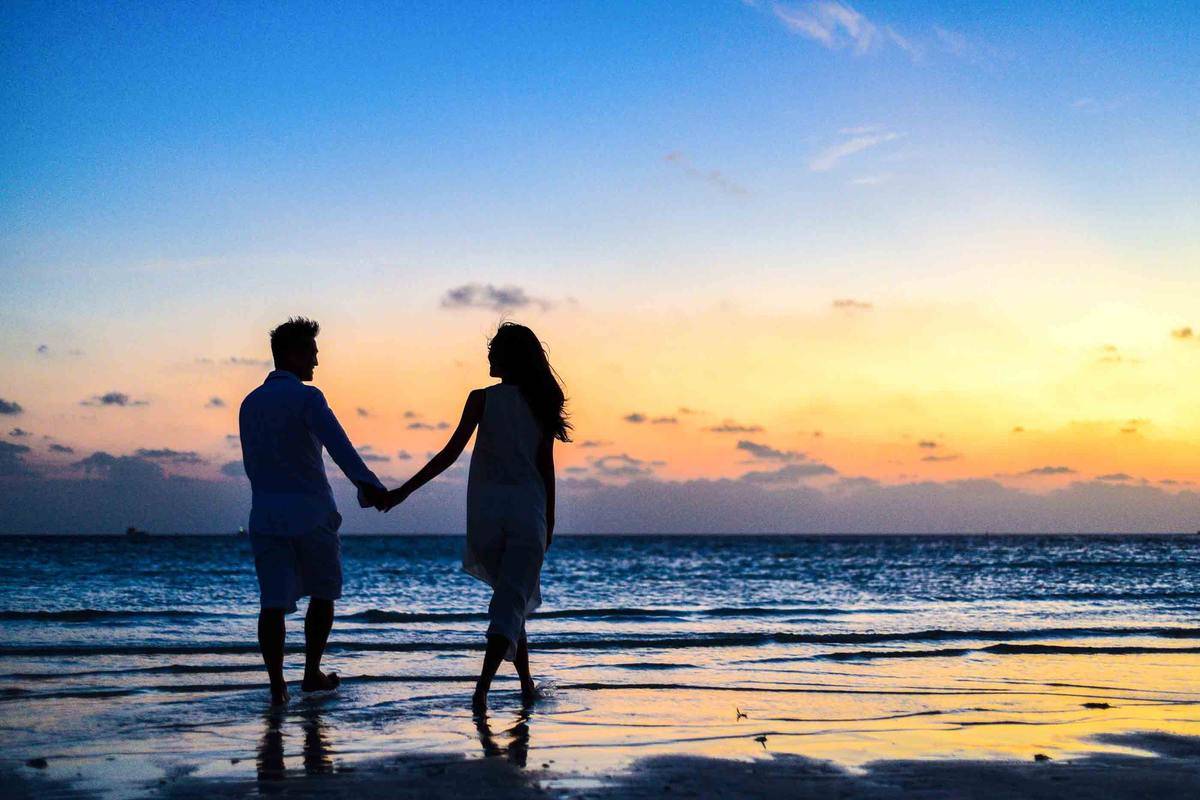 man and woman holding hands walking on beach at sunset