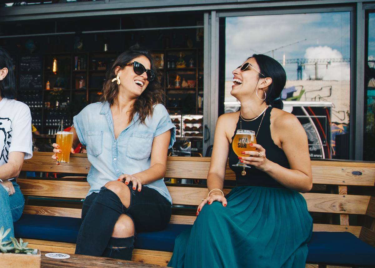two women seated on bench drinking beer laughing