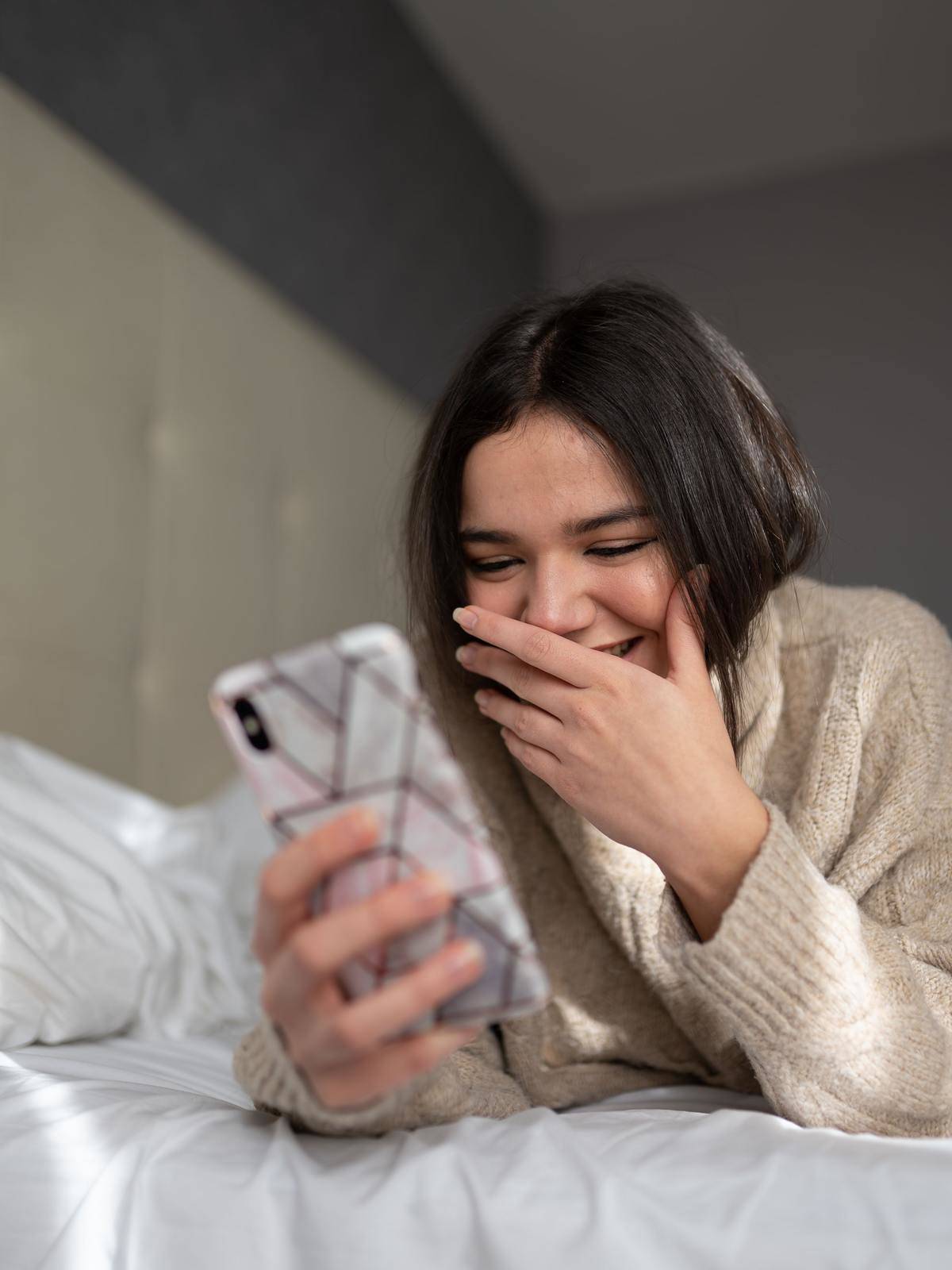 Girl in bed smiling at phone screen and covering mouth