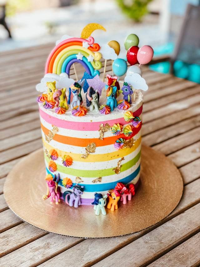 Rainbow cake baked for child's birthday that has rainbows, balloons, and My Little Pony toys