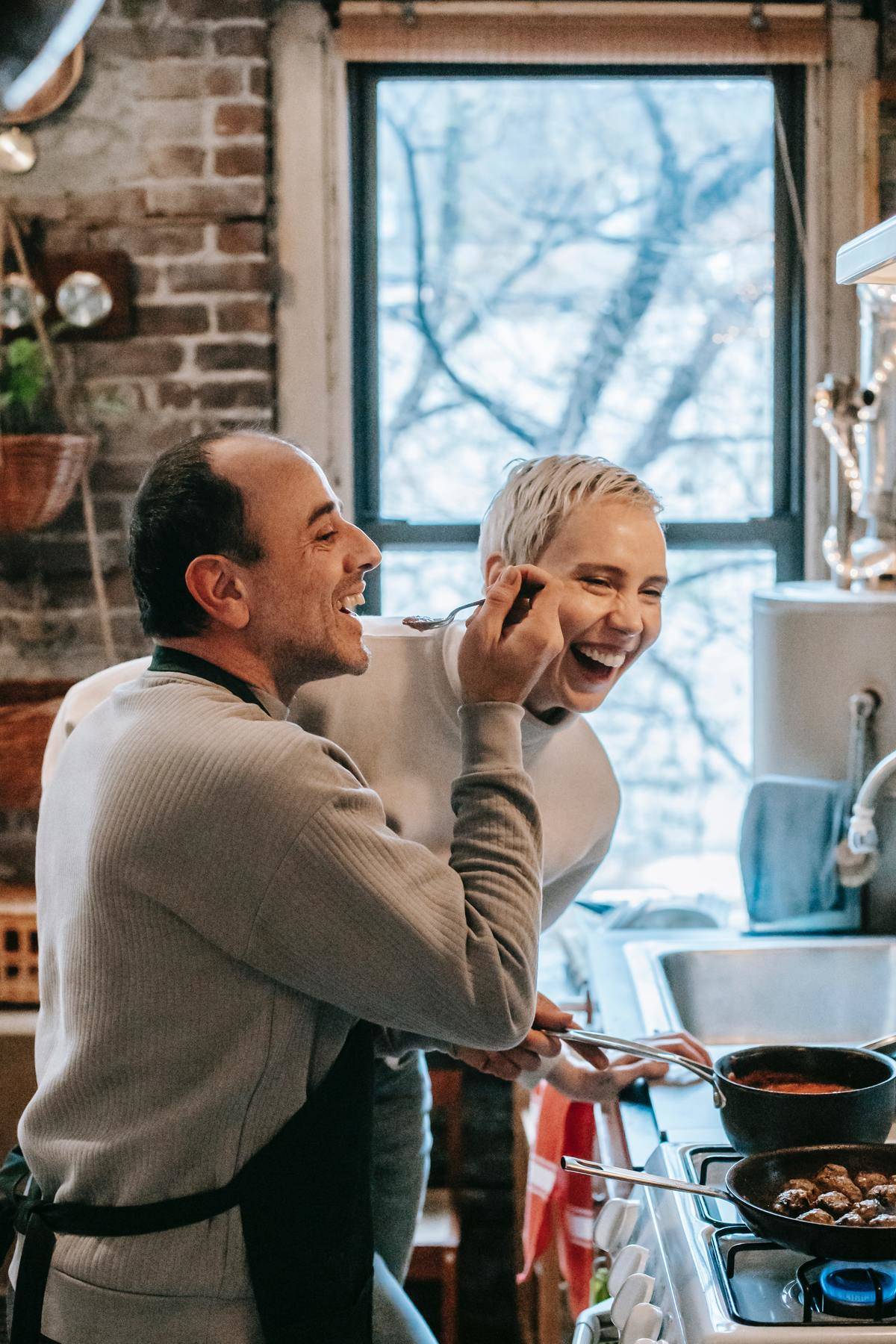 Couple laughs in kitchen, man wears apron and holds spoon for woman to taste