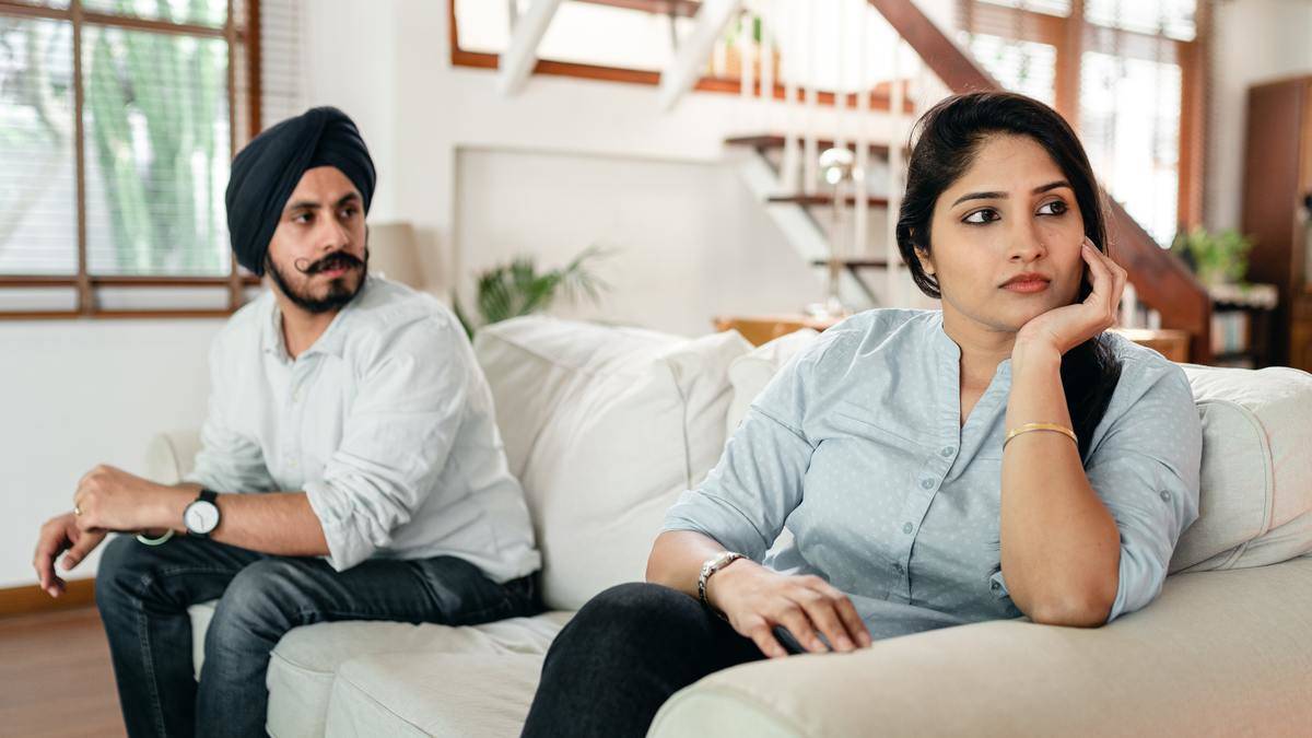 Man and woman sit on couch, woman looks upset, man turned towards her