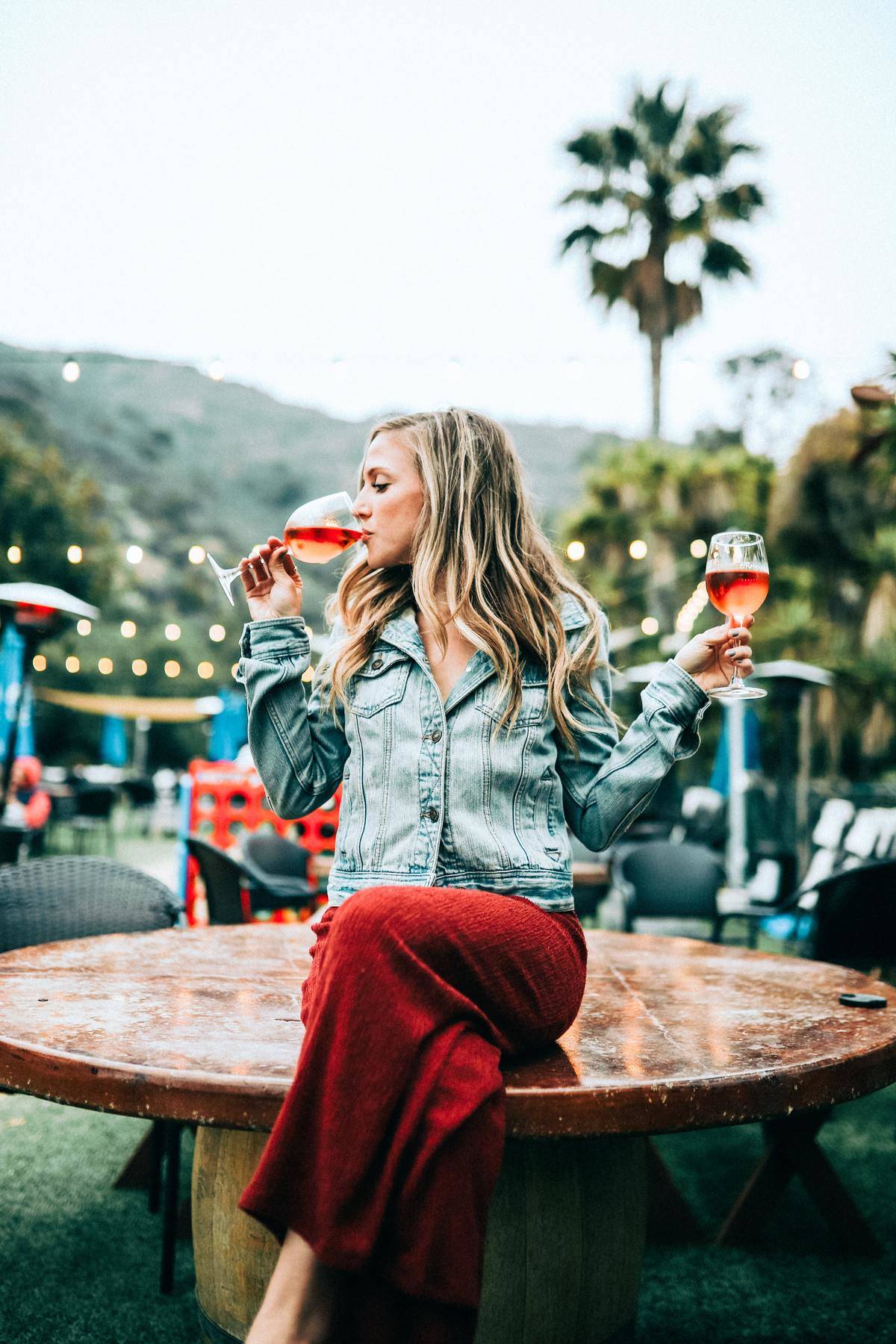 Young blonde woman sits on table with two glasses of wine in hands