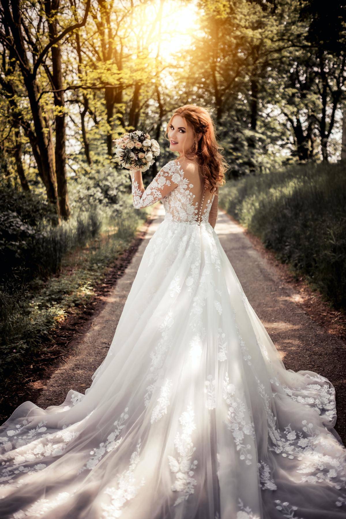 A bride holds her bouquet and walks through a forest at sunset and looks back toward the camera