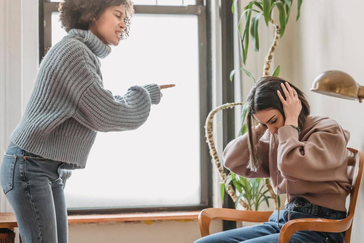 A woman stands up and yells at a woman who is sitting down and covering her ears