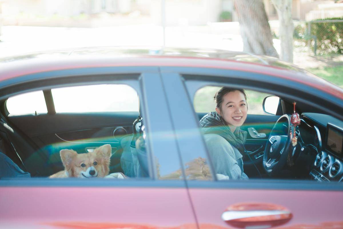 a girl parked in a red car with a corgi in the backseat