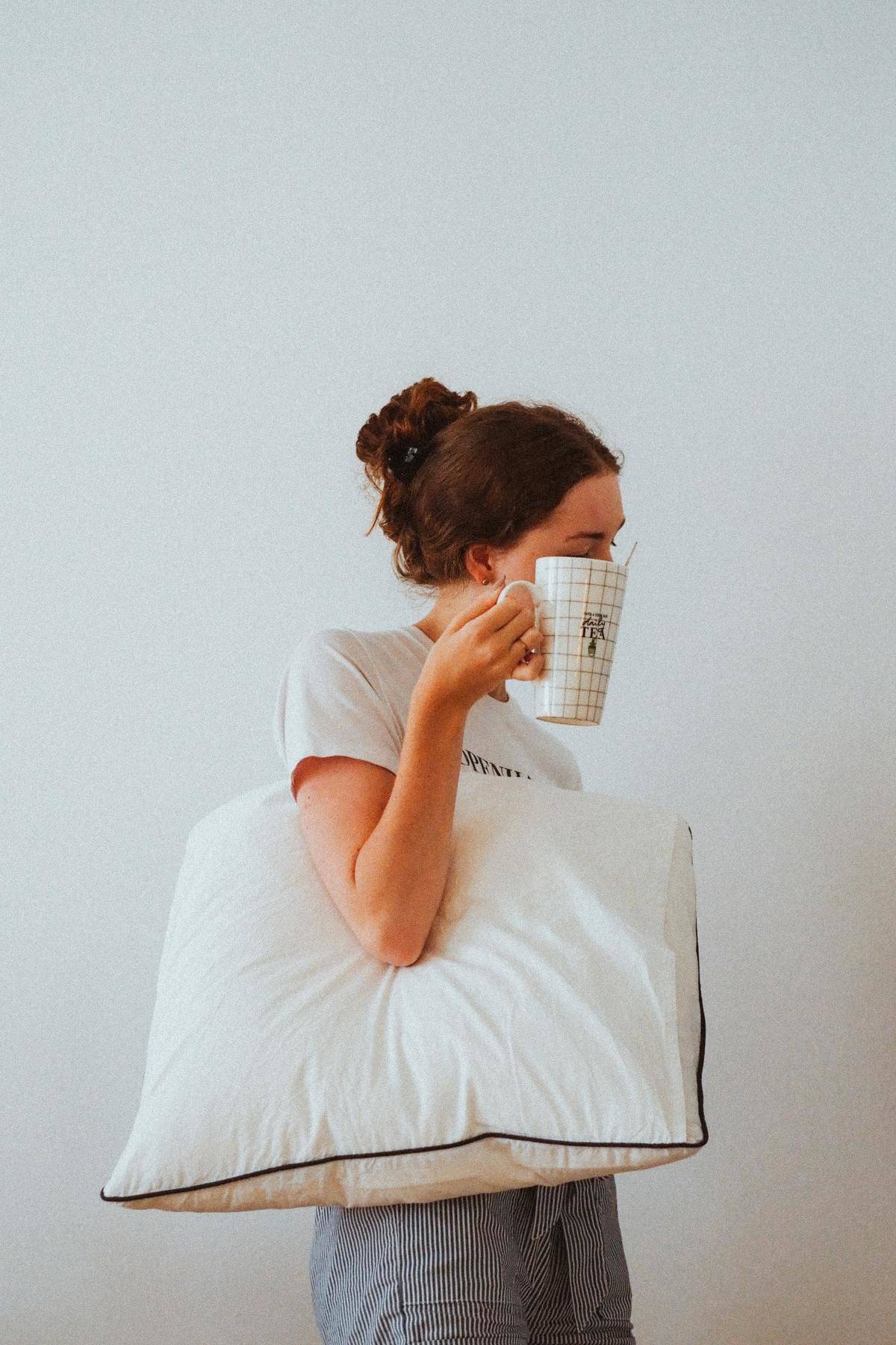 A woman carries a pillow and a tea mug to bed.