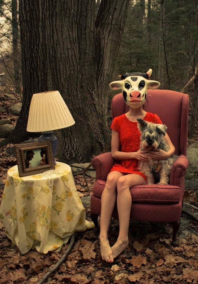 portrait of a seated woman wearing a cow mask in the forest with her dog next to a table with a lamp and framed picture