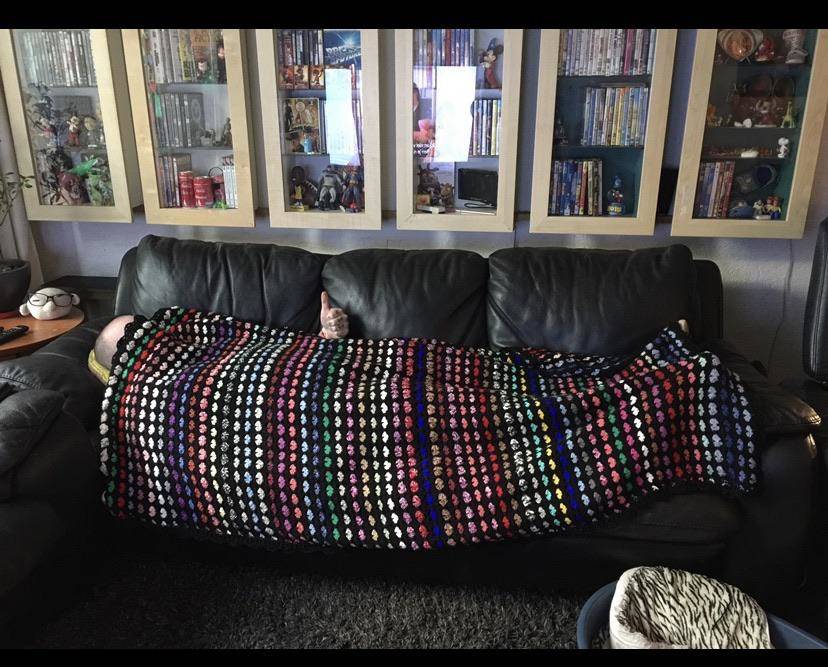 Husband lies under crocheted blanket his wife made for him and gives it a thumbs up.