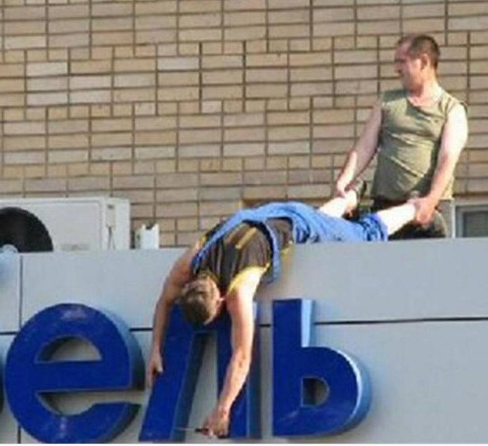 Man hanging off side of a building fixing a sign is being held by his ankles
