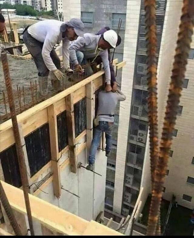Man on side of building being held up by 2 on the roof holding the collar of his shirt