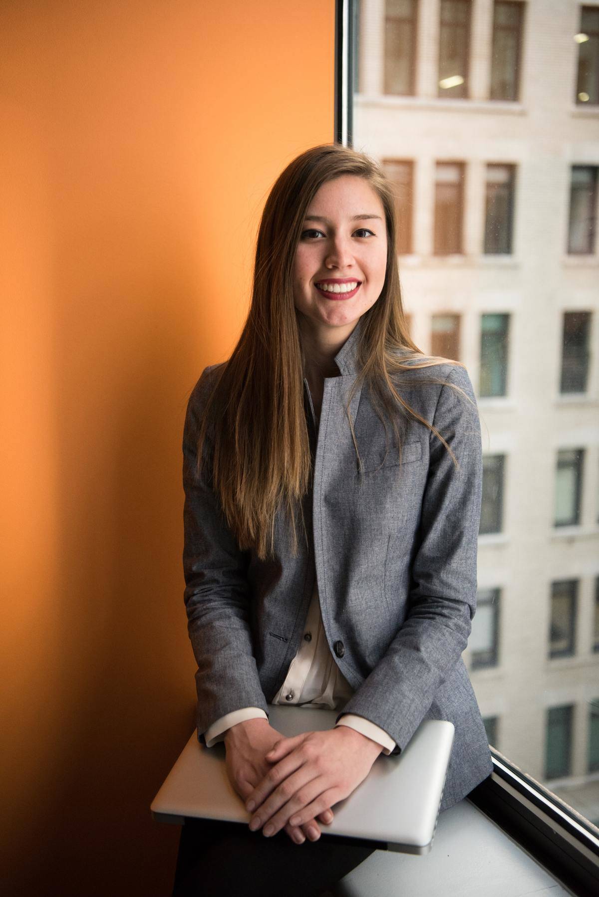 A professional-looking woman sits down and holds a laptop against an orange wall and a building further in the background