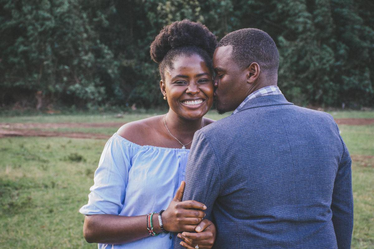 Man and woman hug while she looks at the camera and smiles and he kisses her.
