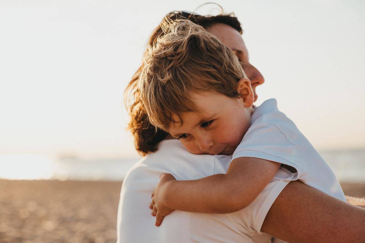 Parent hugs son while the two are outside on the beach.