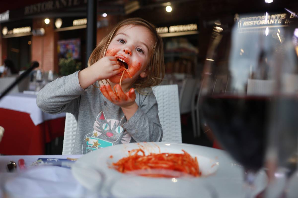 Little girl sits at a table in a restaurant and messily eats pasta.