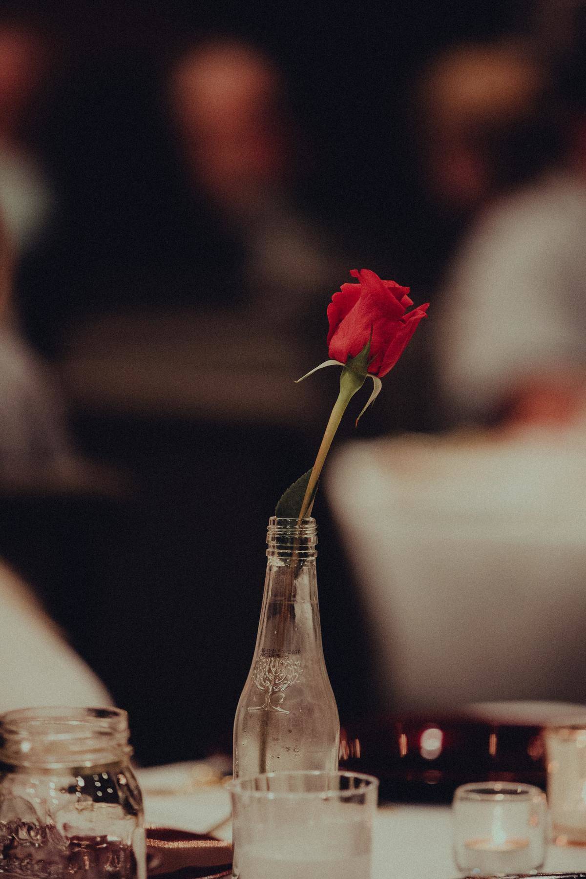 A single red rose sits in a glass bottle on top of a dinner table in a dimly lit restaurant.