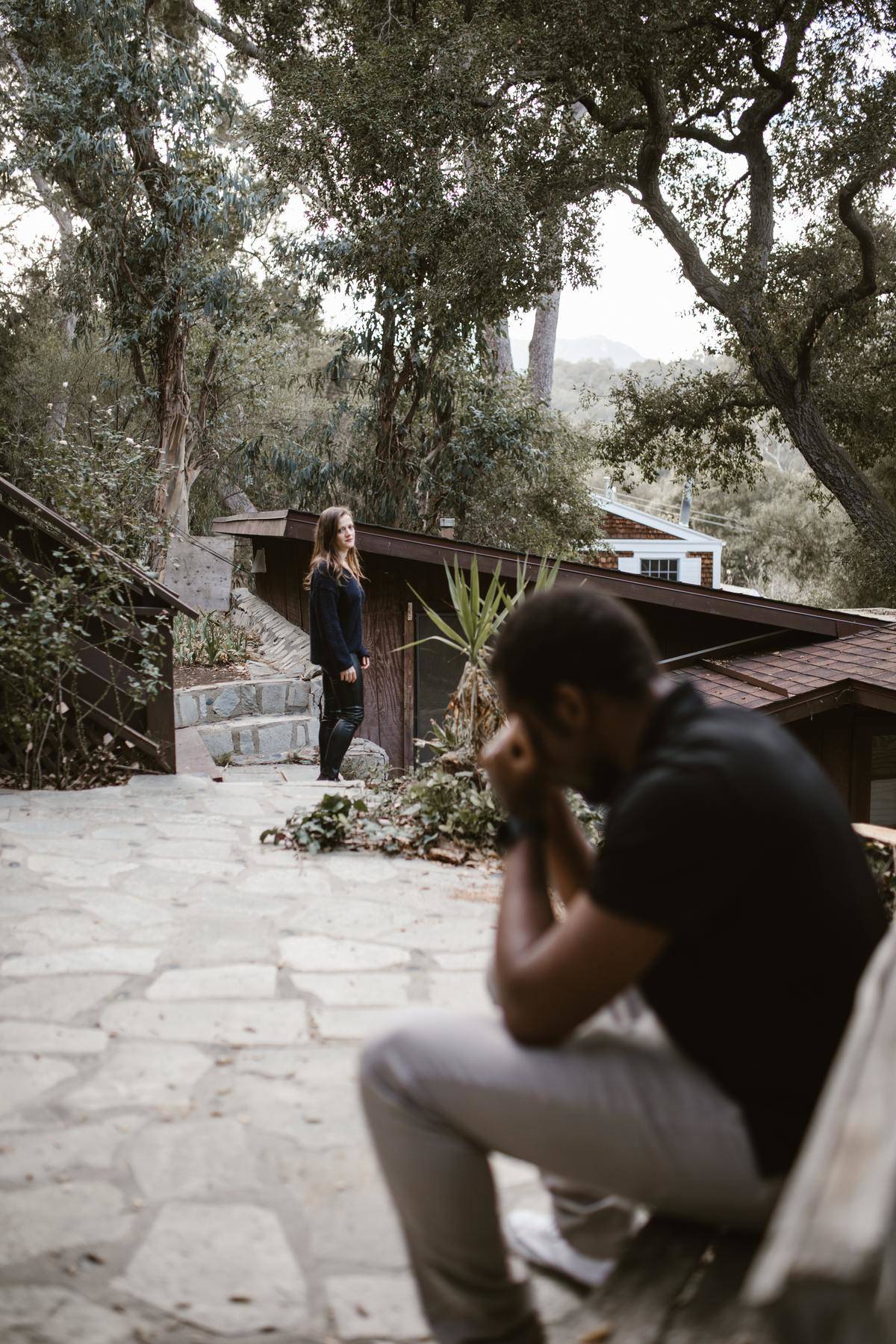 A man sits down on a bench looking upset while a woman stands up further away and looks at him.