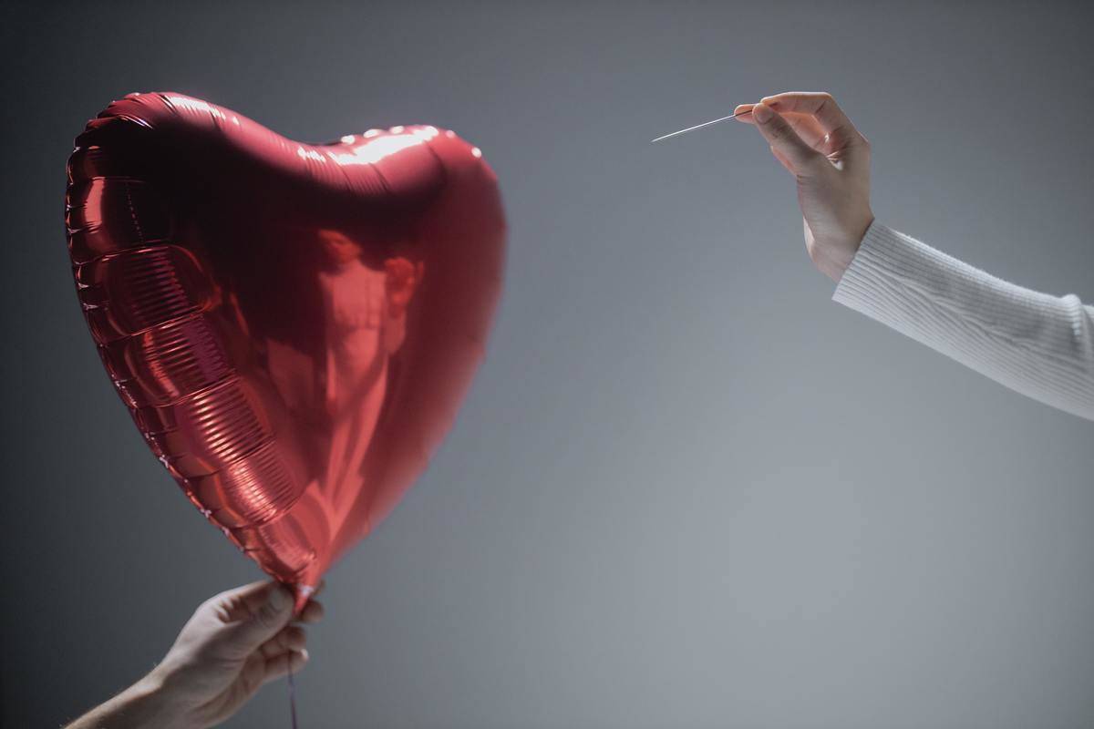 A man holds a heart balloon and a woman approaches it with a needle.
