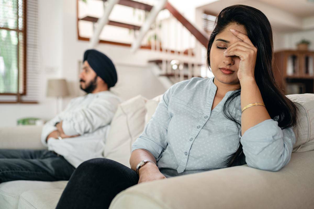 A frustrated wife sits down on the couch next to her husband with her head in her hand.