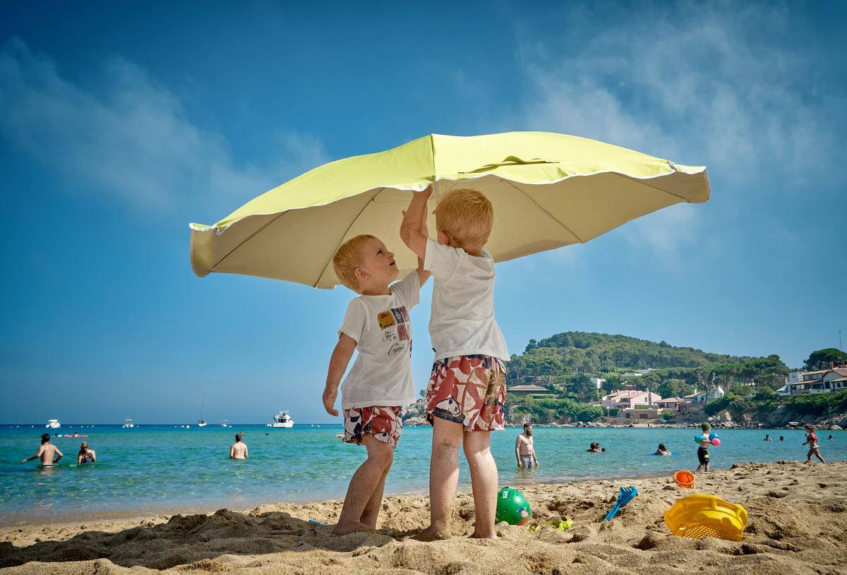 Two boys set up a yellow umbrella on a beach.
