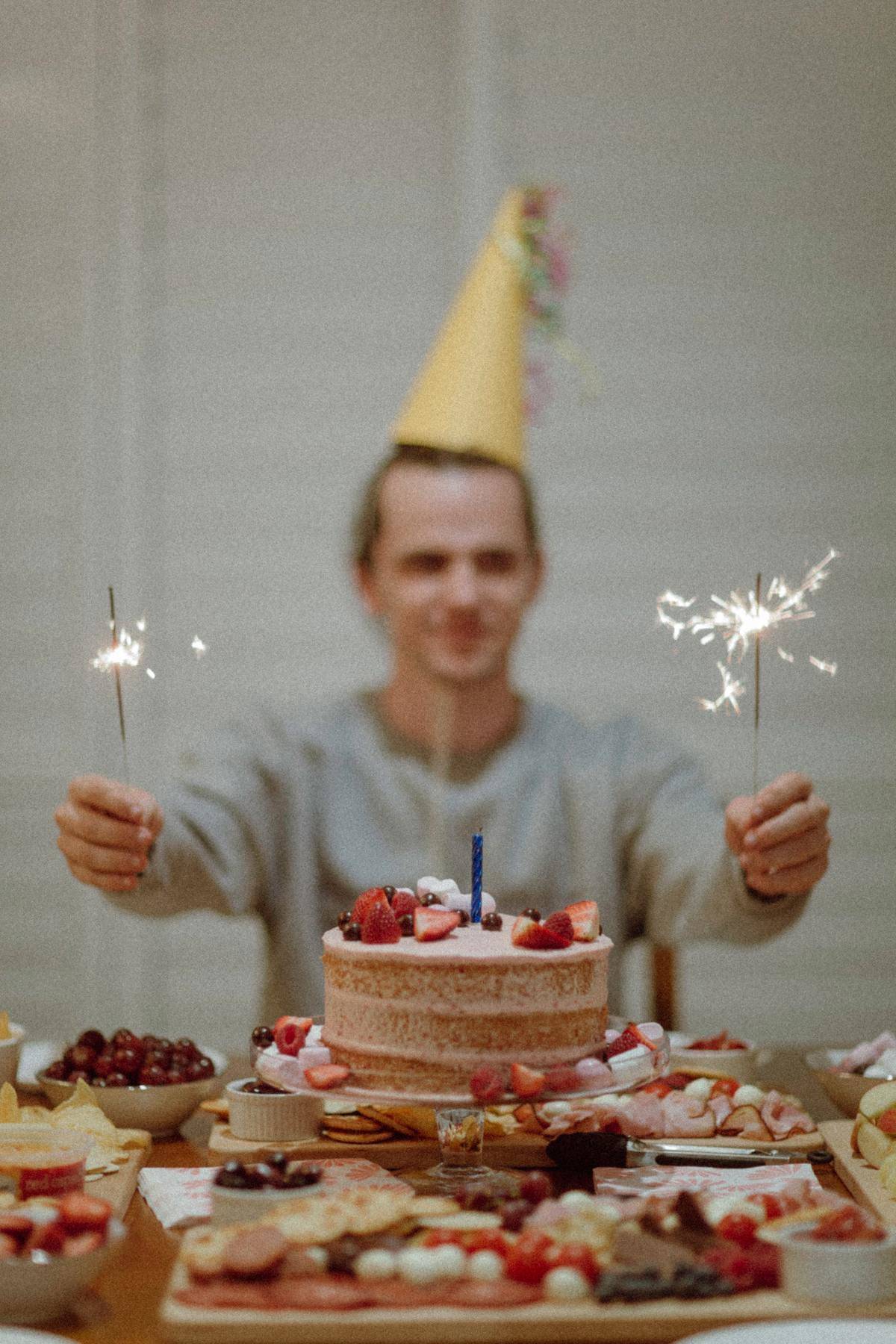 A man celebrating his birthday sits at the head of the table wearing a birthday hat and holding sparklers.