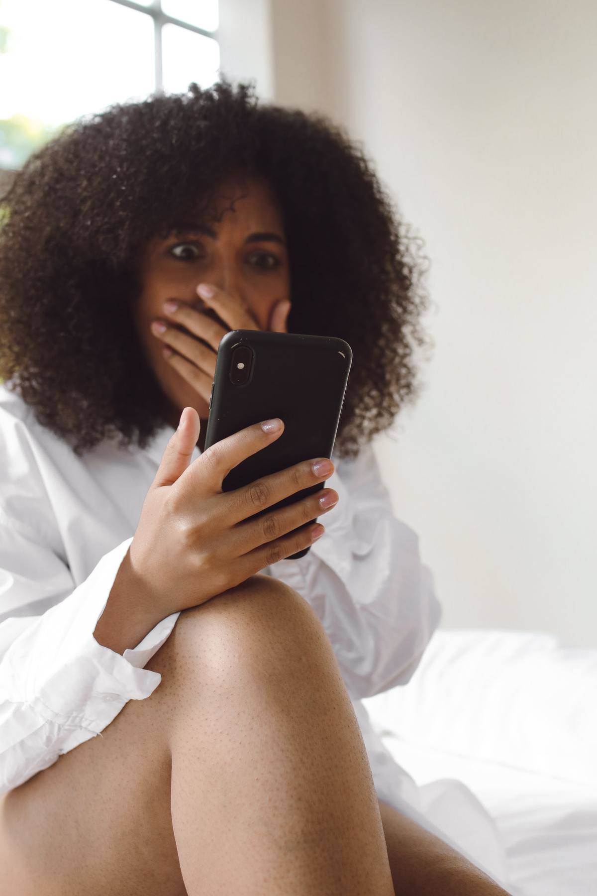 A shocked woman covers her mouth and looks at her phone.