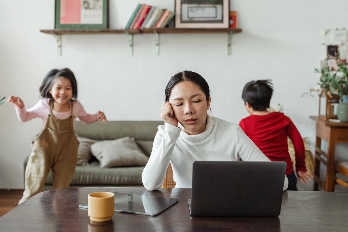 A tired mother works on a laptop while her kids run around behind her.