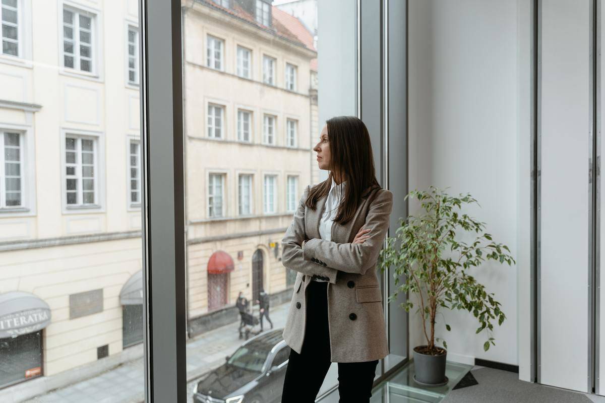 A woman looks out the window of an office with her arms crossed.