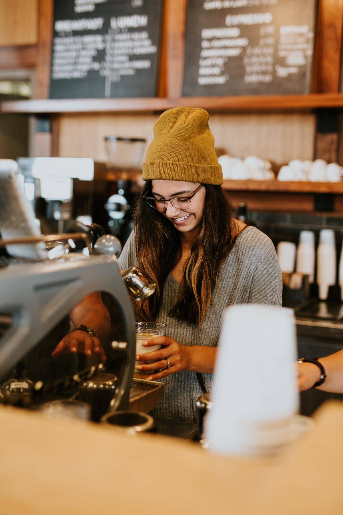 Woman barista wearing yellow beanie smiles while preparing drink