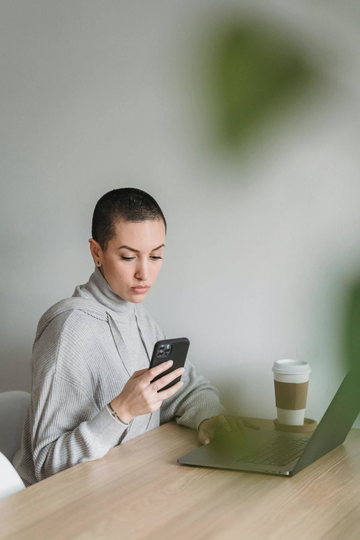 Woman in gray hoodie checks her phone, coffee cup and laptop on table in front of her