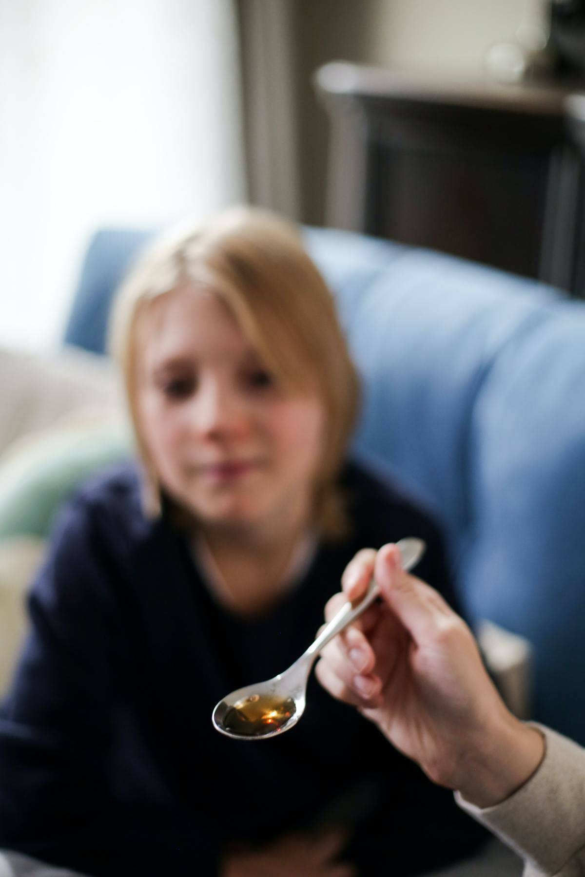 A person holds out a spoon of cough syrup while a child looks at it. 