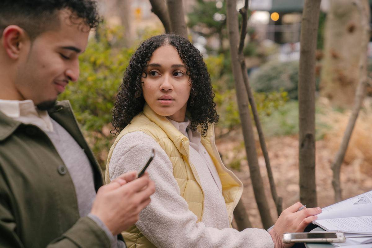 Man and woman sitting on park bench. Man is on his phone and woman is looking at him
