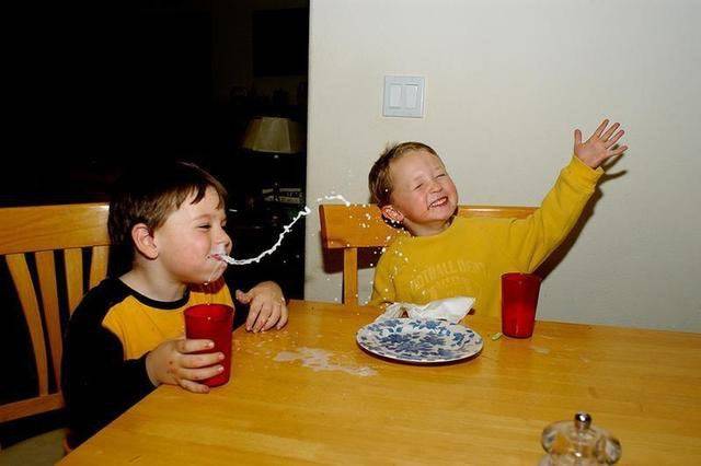 Two kids at a table and one is spitting milk out of their mouth.