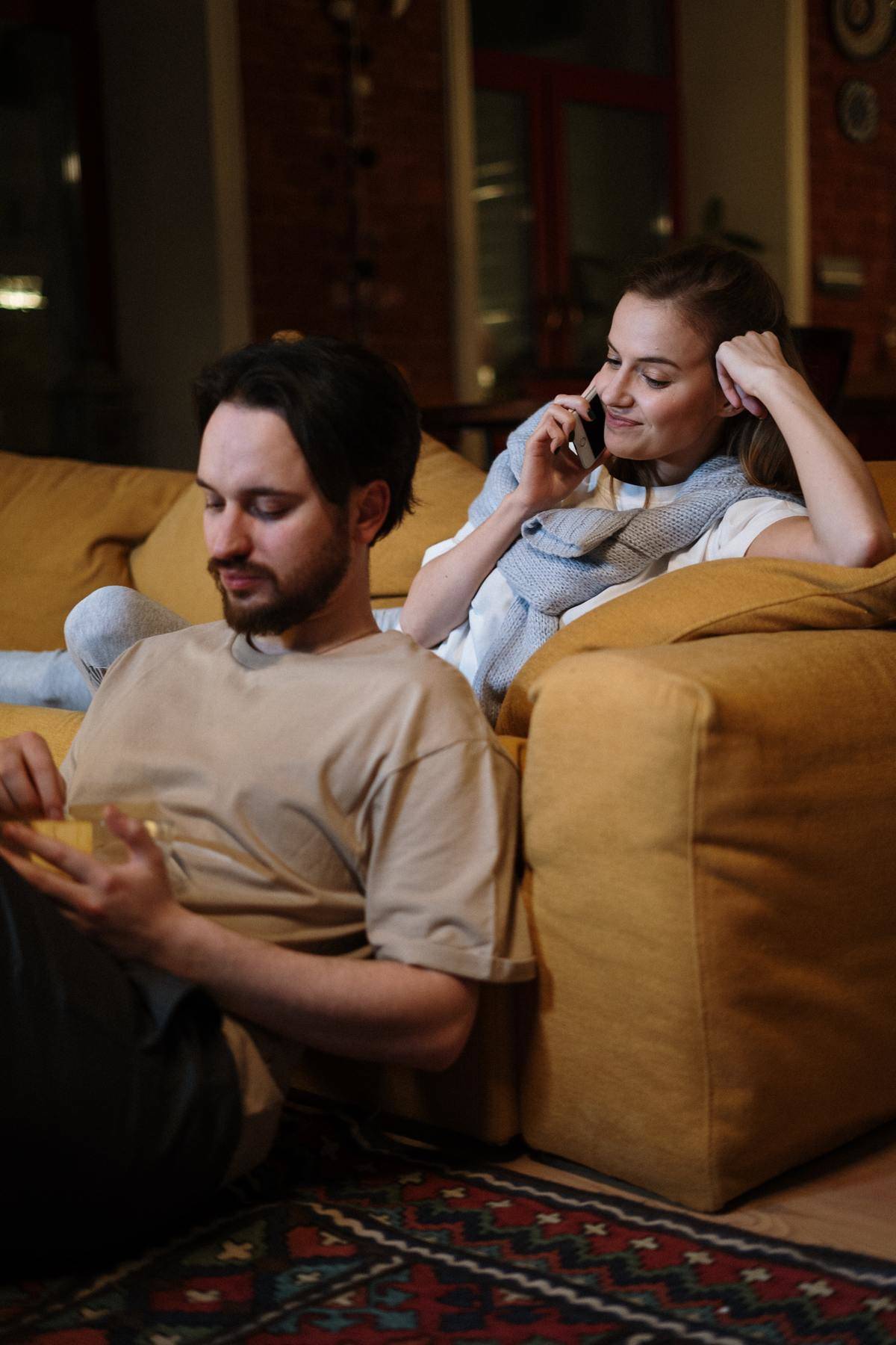 Man and woman in living room. Woman is sitting on the couch talking on the phone. Man is sitting on the floor