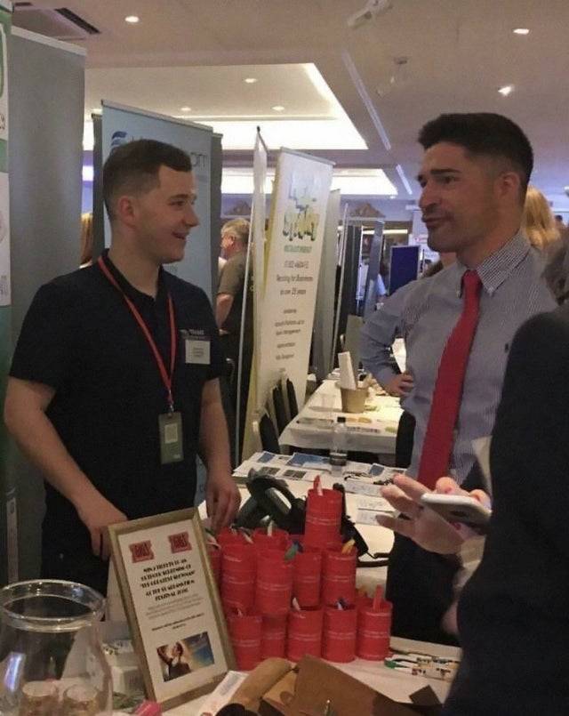 Two men talking at a table and one looks to have a very small arm.