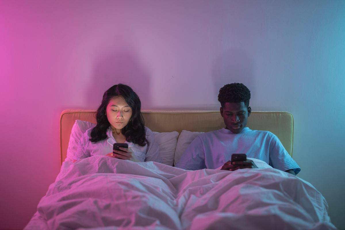 Couple in bed in pink lighting, both looking at their cell phones