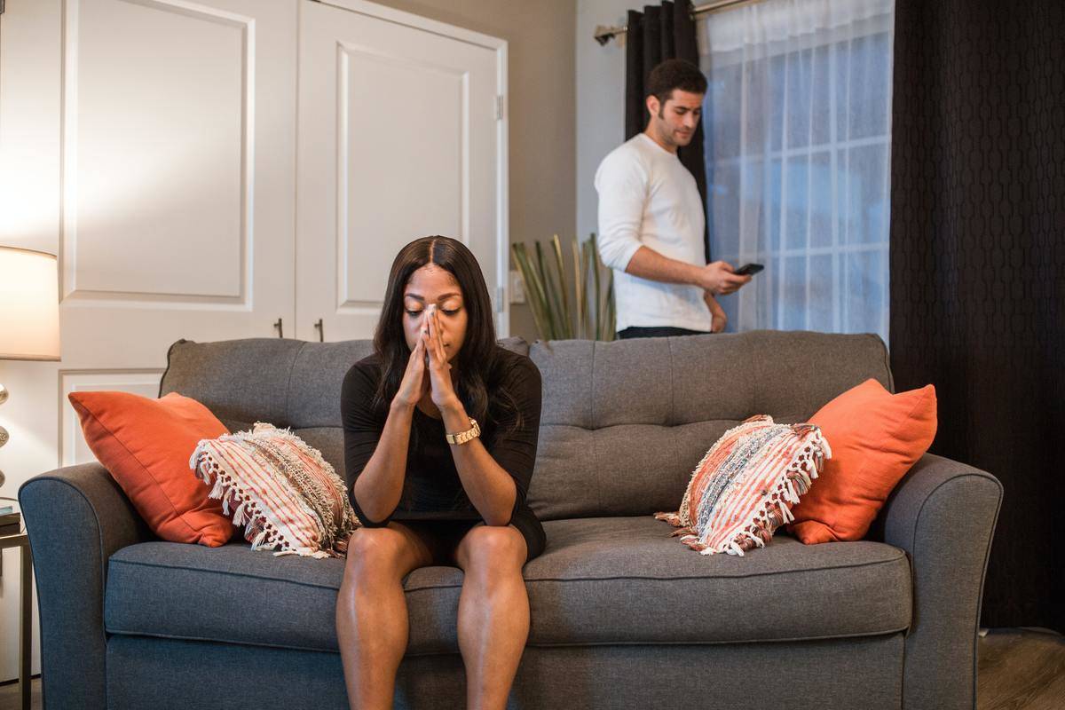 Man looking at phone while woman sits on couch looking stressed