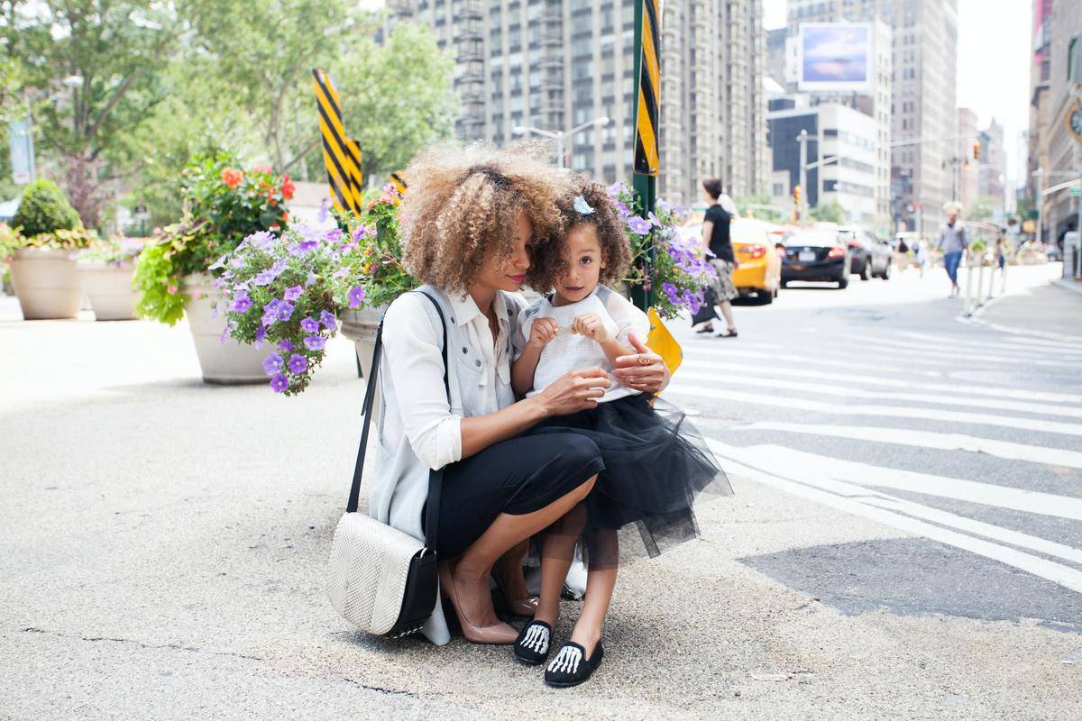 A mother holds her daughter while they stand outside on a city sidewalk.