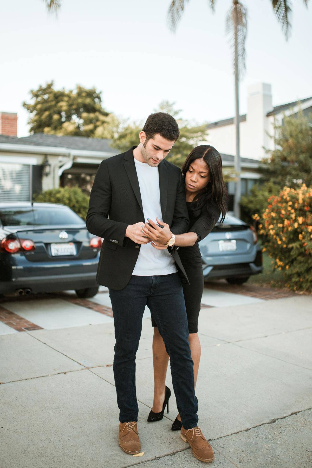 Woman leaning over man's shoulder looking at his phone, both wear black 
