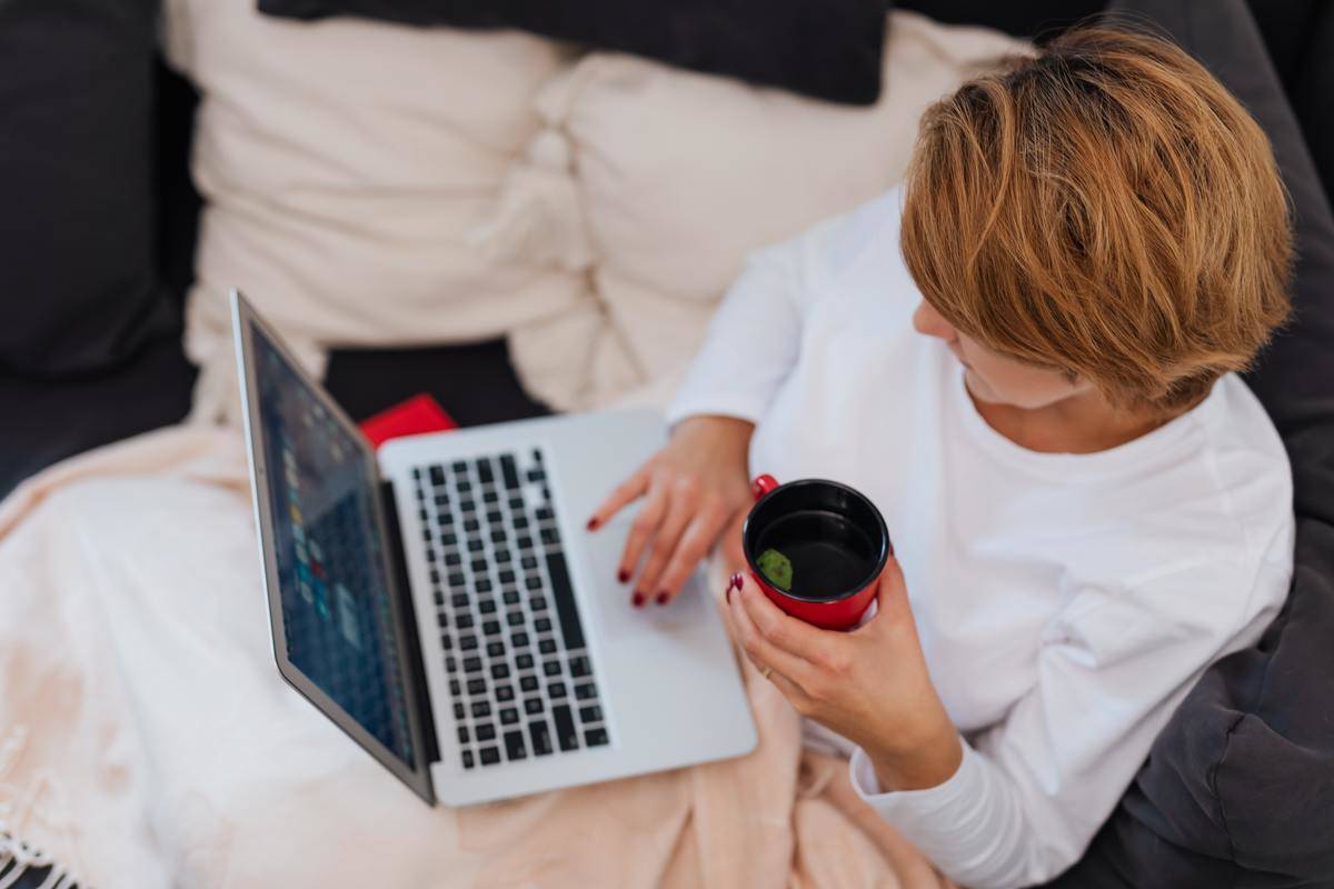 Woman sitting on couch with blanket holding tea typing on laptop 