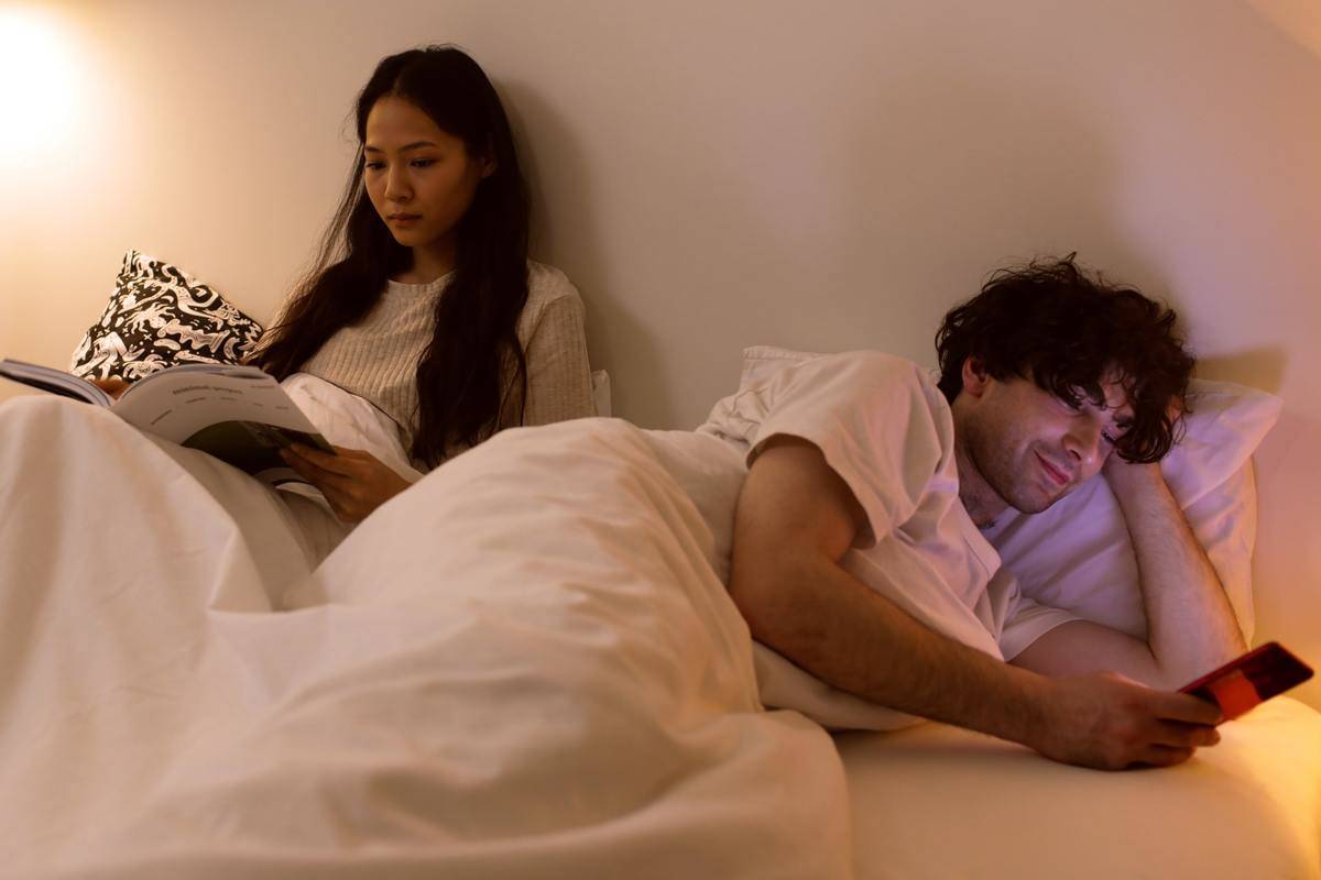 Woman in bed reading, man rolled over beside her smiling at cell phone