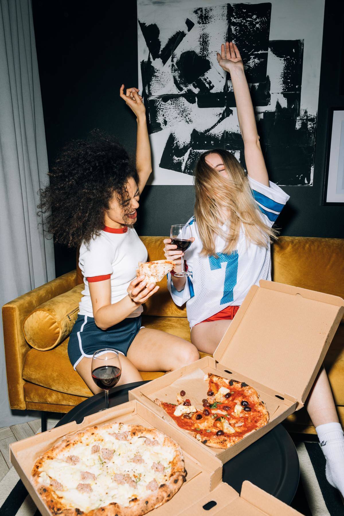 Women partying on couch with open pizza box and wine